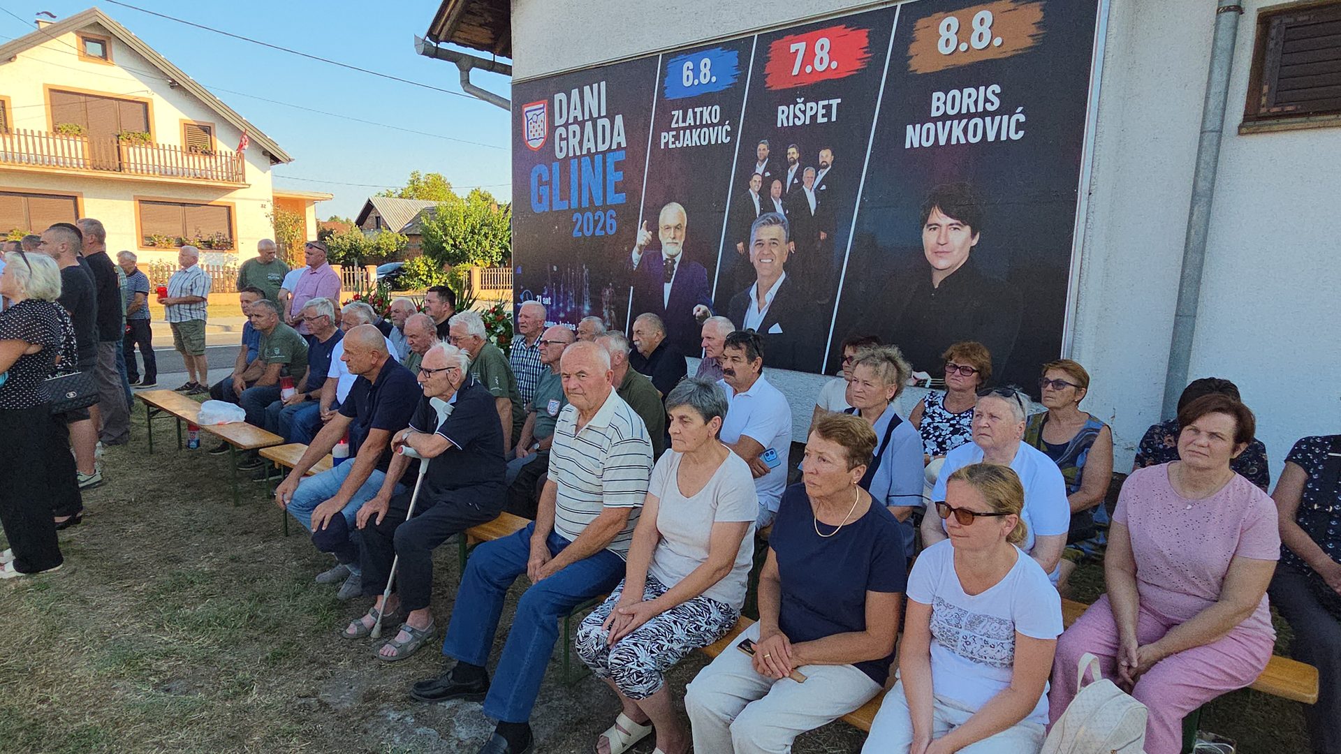 Skupina ljudi sjedi na otvorenome na drvenim klupama ispred zida zgrade na kojem visi veliki plakat s najavom koncerata.