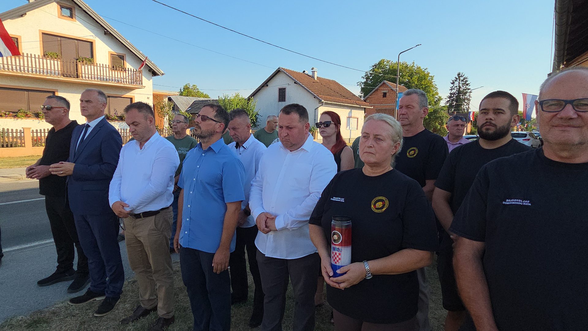 Ljudi u mirnom stavu stoje na cestovnom proširenju ispred kuća, dok žena u prvom planu drži lampion s hrvatskim grbom.