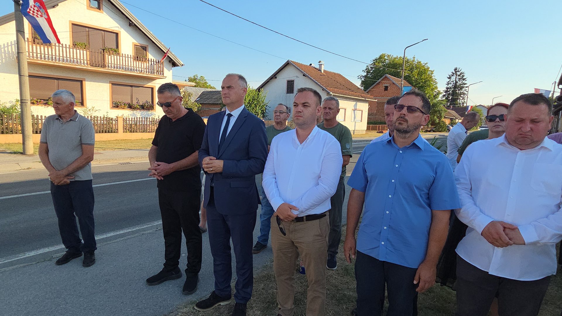 Muškarci u odijelima i košuljama stoje uz cestu s rukama sklopljenim ispred sebe u znak poštovanja.