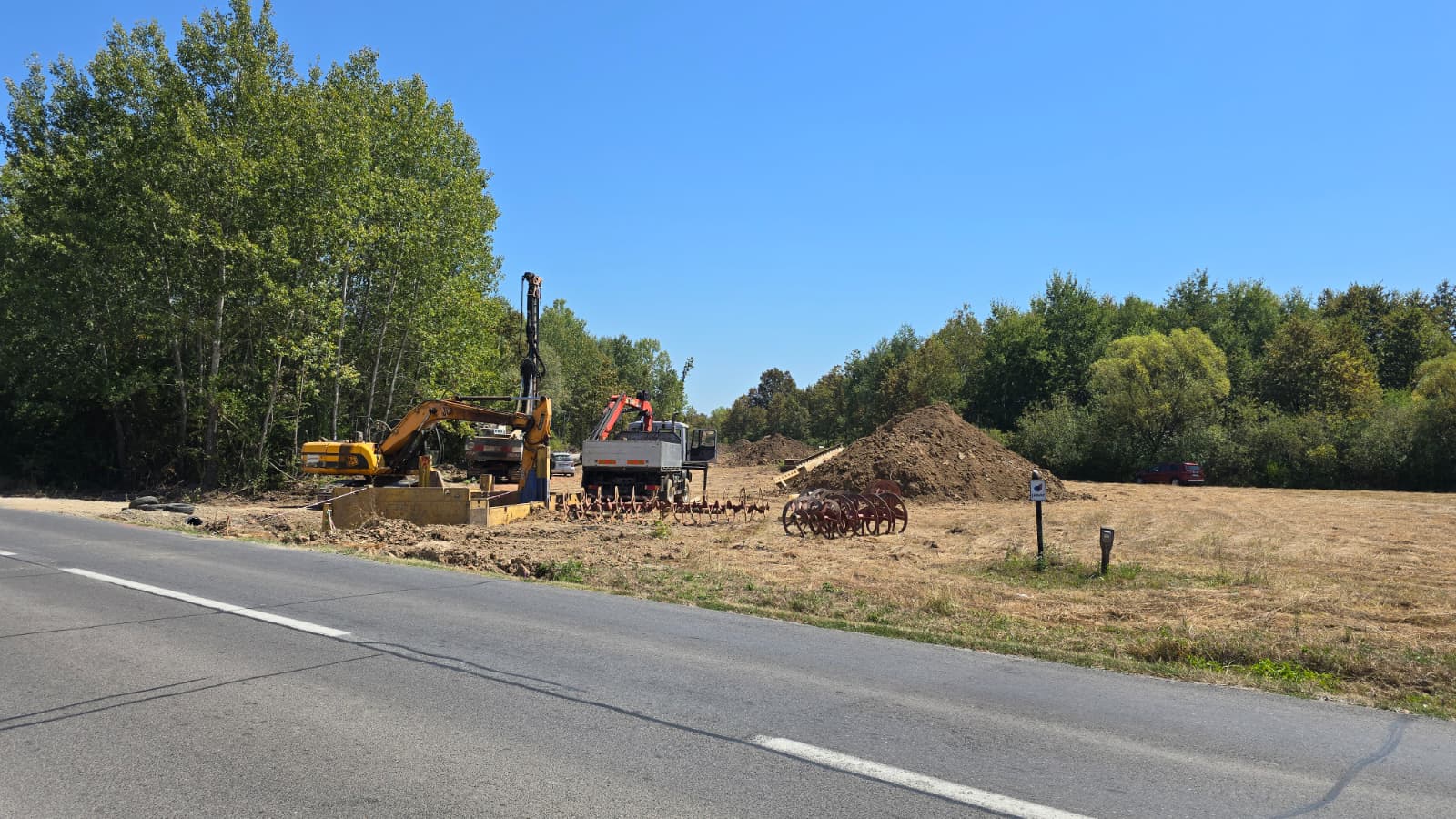 Žuti bager, kamion i hrpa zemlje na gradilištu pokraj asfaltirane ceste i šume.