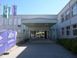 Prilaz i natkriveni ulaz u svijetlosivu zgradu uz građevinsku ogradu i zastave.