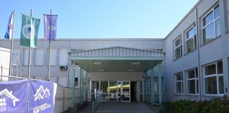 Prilaz i natkriveni ulaz u svijetlosivu zgradu uz građevinsku ogradu i zastave.