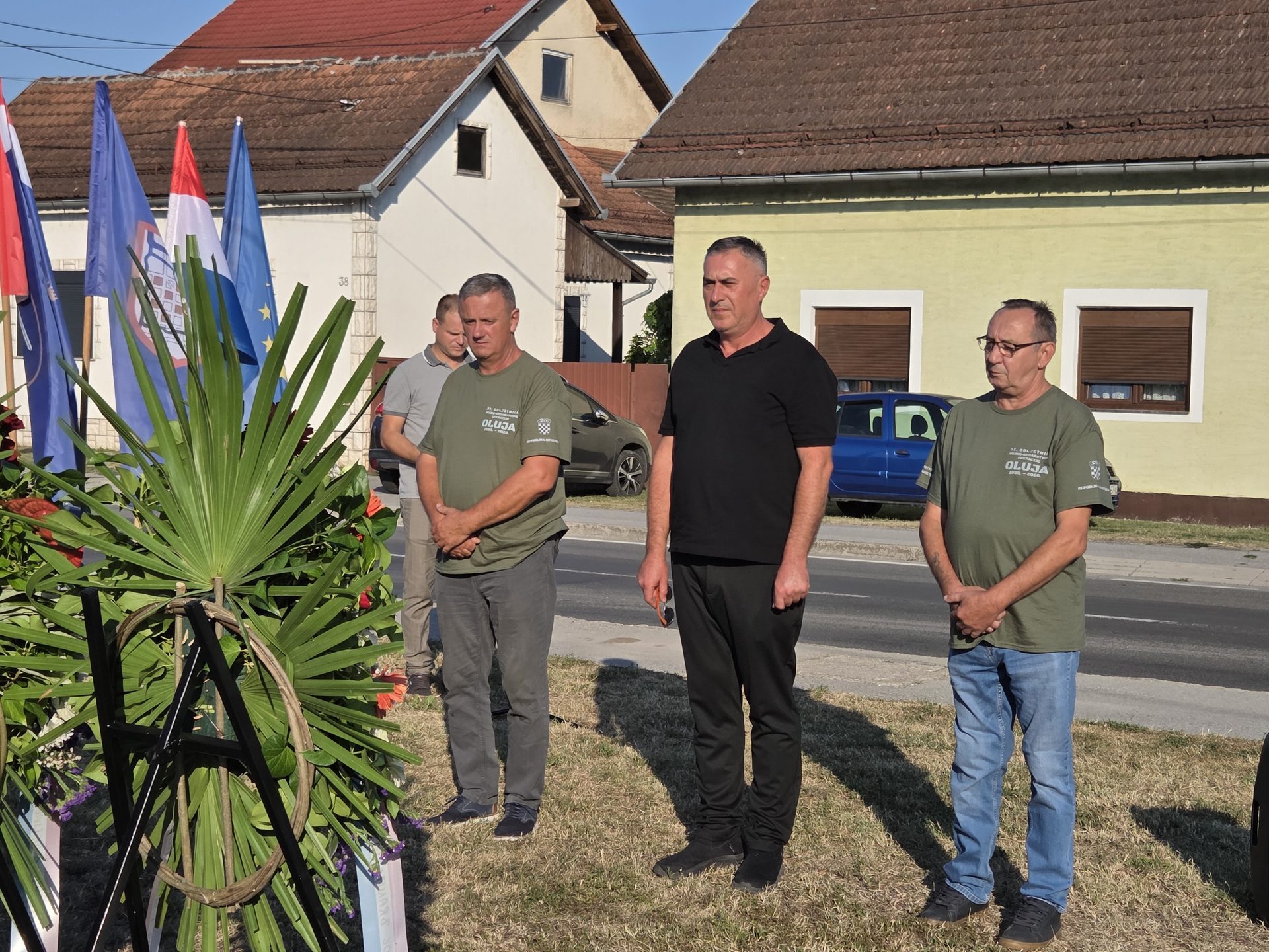 Tri muškarca stoje mirno pokraj cvjetnog vijenca i postavljenih zastava ispred obiteljskih kuća uz cestu.