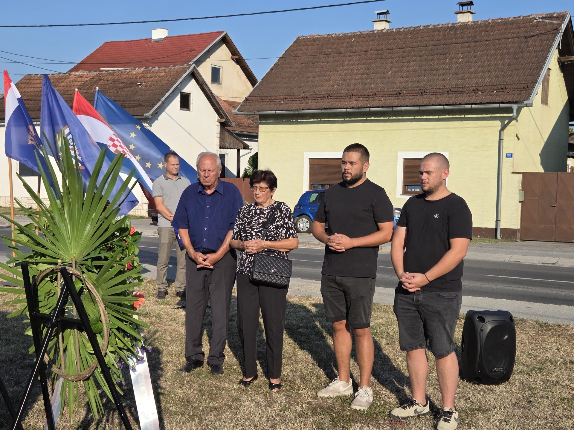 Skupina ljudi stoji u tišini uz cvjetni vijenac i hrvatske te europske zastave pokraj ceste.